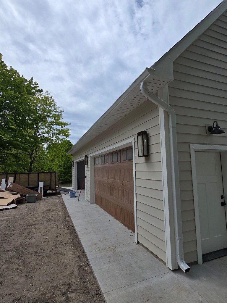 **Image Description:** * A garage with a brown door and white siding, featuring two white downspouts for rainwater drainag...