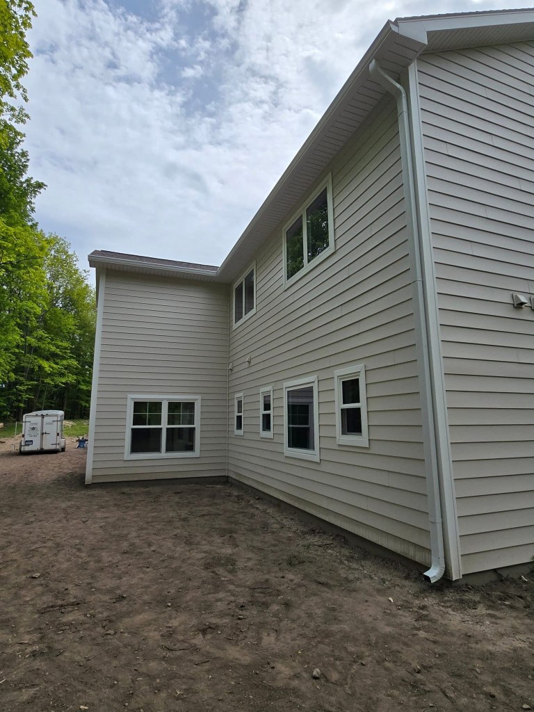 The house has the color of tan vinyl siding, white-colored trim and roofing and white-colored gutters. The backyard of the...